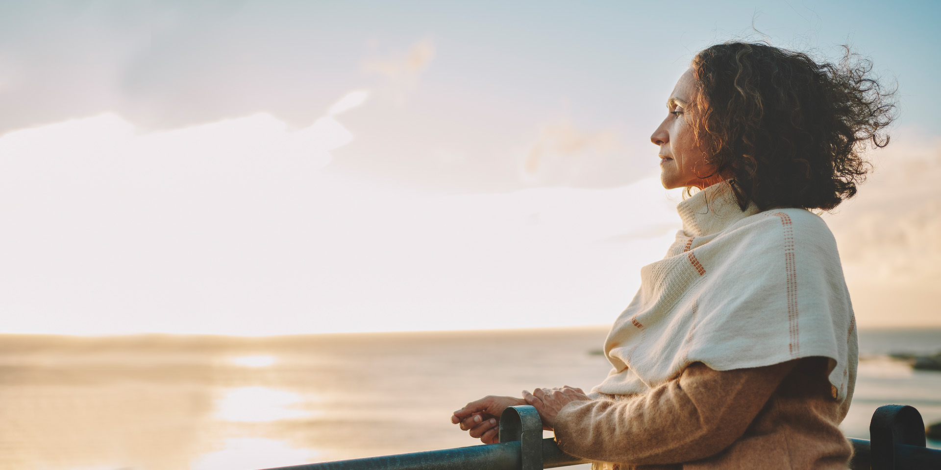 A woman stands against a railing looking out over the horizon towards the sun with a look of quiet confidence.